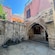 An ancient stone fountain with spouts dripping water next to an arched entryway surrounded by buildings